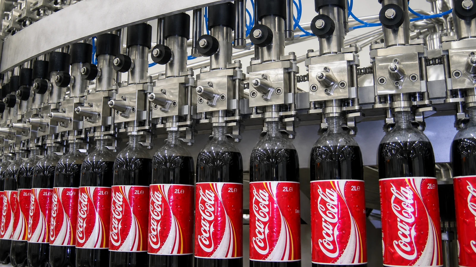 Coca-Cola PET bottles with red labels beneath stainless-steel filling valves on a carbonated soft drink bottling line